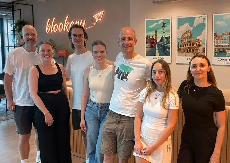 Das blookery-Team steht vor dem Firmenlogo und Städteplakaten im Büro
