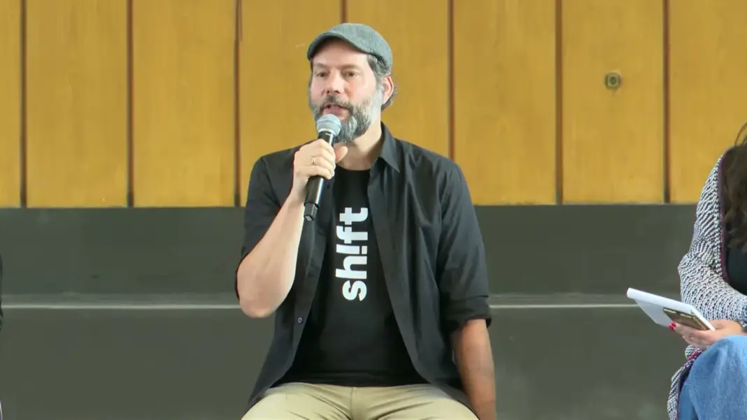Carsten Waldeck mit grauer Kappe und schwarzem Shirt mit dem Schriftzug shift spricht im Sitzen ins Mikrofon