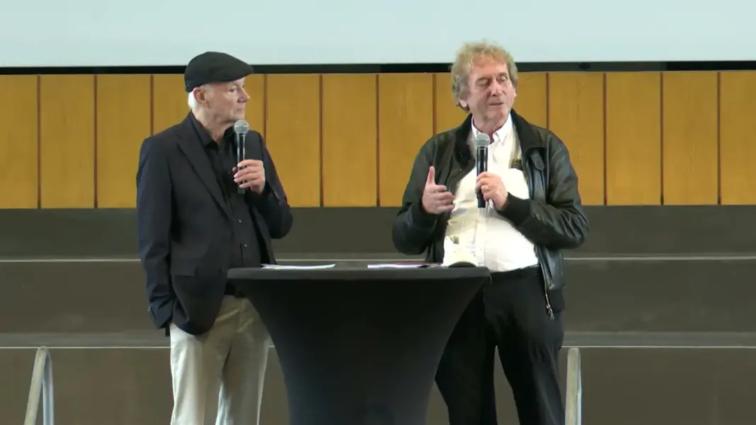 Günter Faltin mit Mütze und Michael Braungart stehen mit Mikrofonen an einem Stehtisch auf der Bühne des Audimax