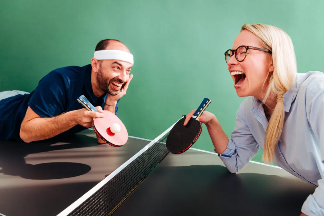 Zwei lachende Spieler mit Tischtennisschlaegern beugen sich von beiden Seiten ueber das Netz einer Platte