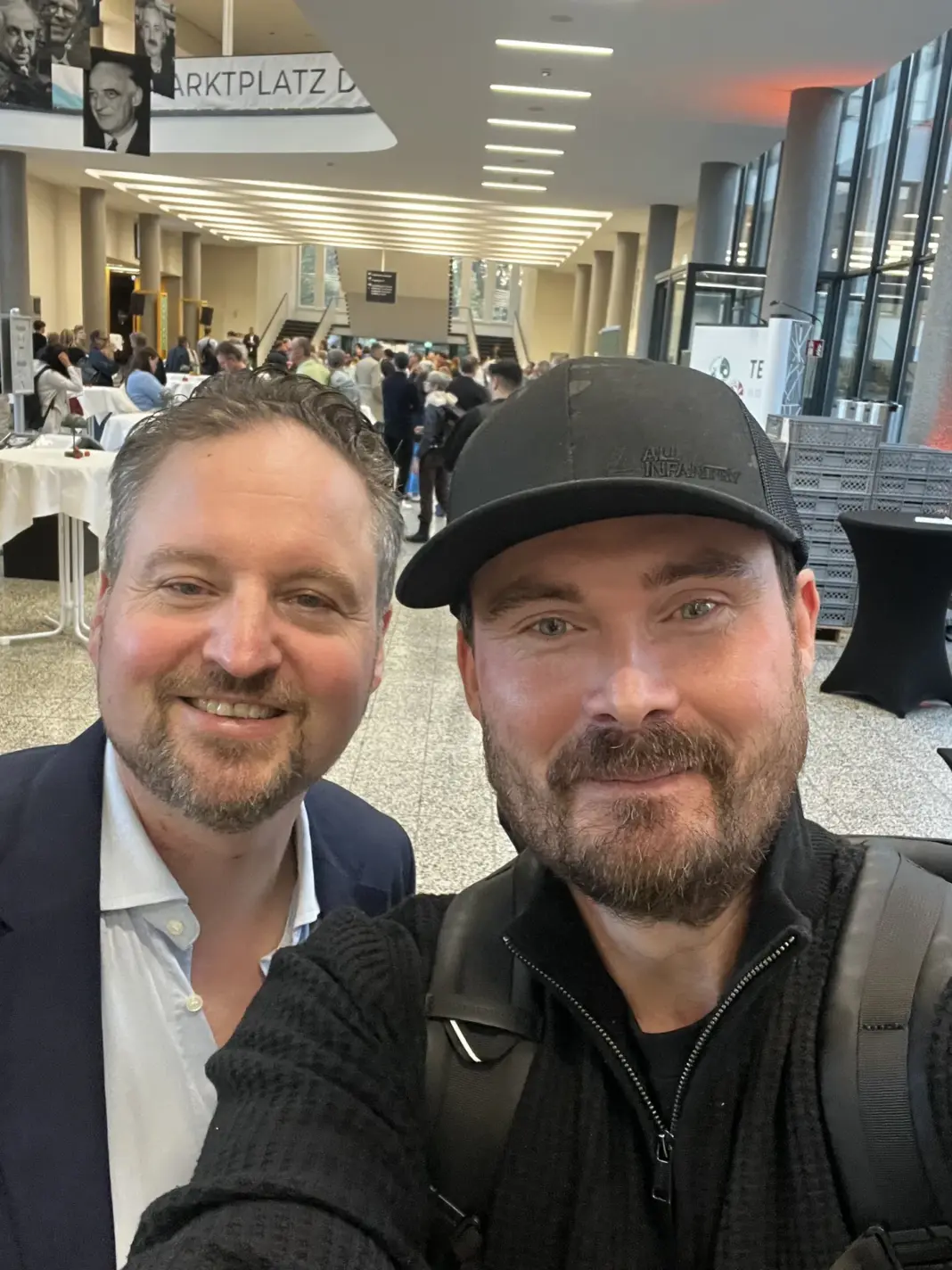 Dennis Weidner von Gründerfreunde und Florian Komm von der Stiftung Entrepreneurship lächeln in die Kamera, im Hintergrund das volle Foyer des Henry-Ford-Baus