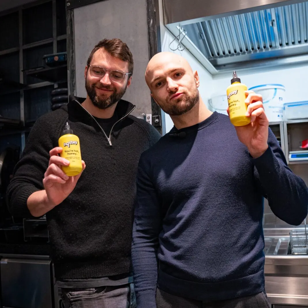 Die beiden Hottey-Gründer halten in einer Küche je eine Flasche Hottey in die Kamera