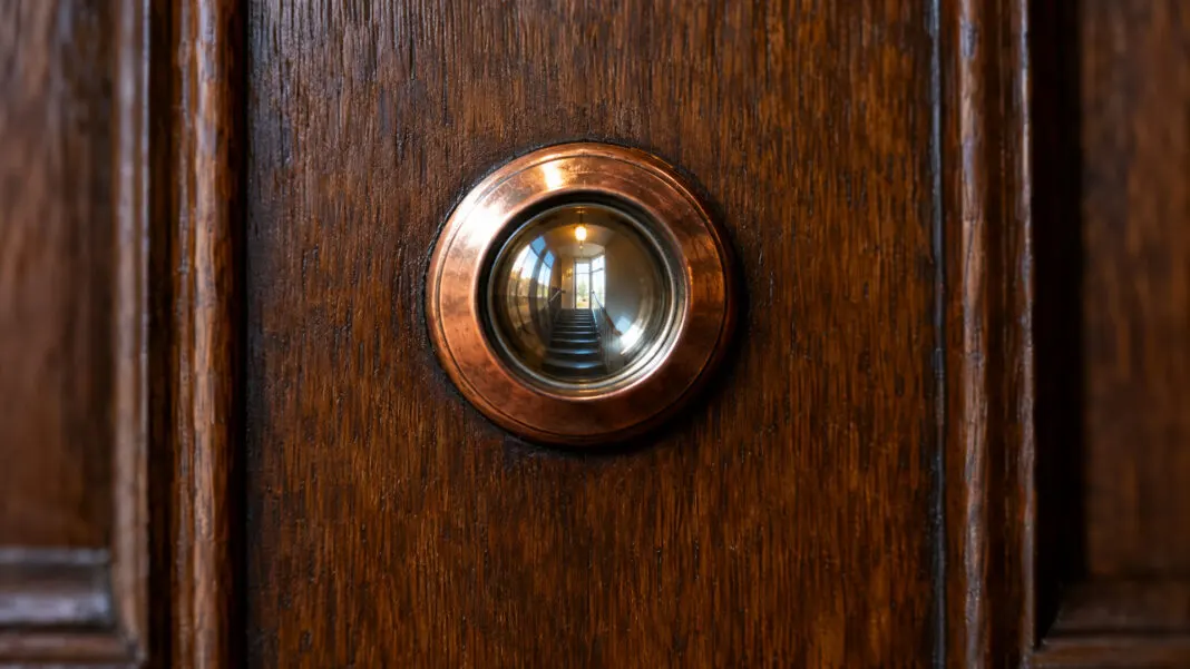 Kupferner Türspion in einer geschlossenen Eichentür, das Glas spiegelt das Licht des Treppenhauses