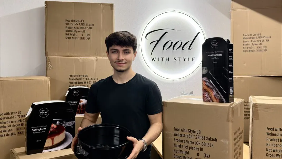 Emre Tuncer hält eine schwarze Springform, um ihn stehen Versandkartons und verpackte Backformen von Food with Style vor dem beleuchteten Firmenschild