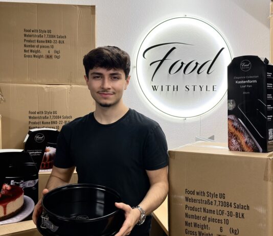 Emre Tuncer hält eine schwarze Springform, um ihn stehen Versandkartons und verpackte Backformen von Food with Style vor dem beleuchteten Firmenschild