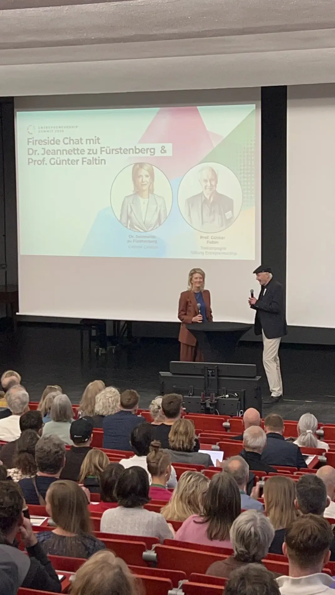 Jeannette zu Fürstenberg im braunen Anzug und Günter Faltin mit Mikrofon auf der Bühne vor der Leinwand mit ihren Porträts, davor das Publikum