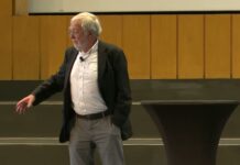 Neurobiologe Gerald Hüther im dunklen Sakko spricht frei auf der Bühne im Audimax der FU Berlin und zeigt mit der Hand in den Saal