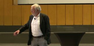 Neurobiologe Gerald Hüther im dunklen Sakko spricht frei auf der Bühne im Audimax der FU Berlin und zeigt mit der Hand in den Saal