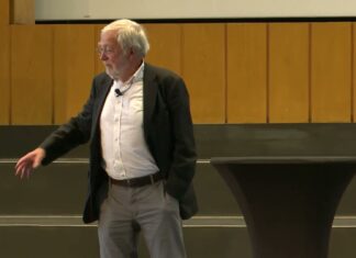 Neurobiologe Gerald Hüther im dunklen Sakko spricht frei auf der Bühne im Audimax der FU Berlin und zeigt mit der Hand in den Saal