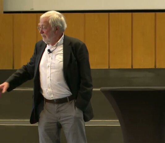 Neurobiologe Gerald Hüther im dunklen Sakko spricht frei auf der Bühne im Audimax der FU Berlin und zeigt mit der Hand in den Saal
