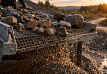 Ein grobes Drahtsieb aus verzinktem Stahl trennt feinen Kies von faustgroßen Feldsteinen, die Körner rieseln durch das Gitter, schräges Abendlicht auf nassem Stein.