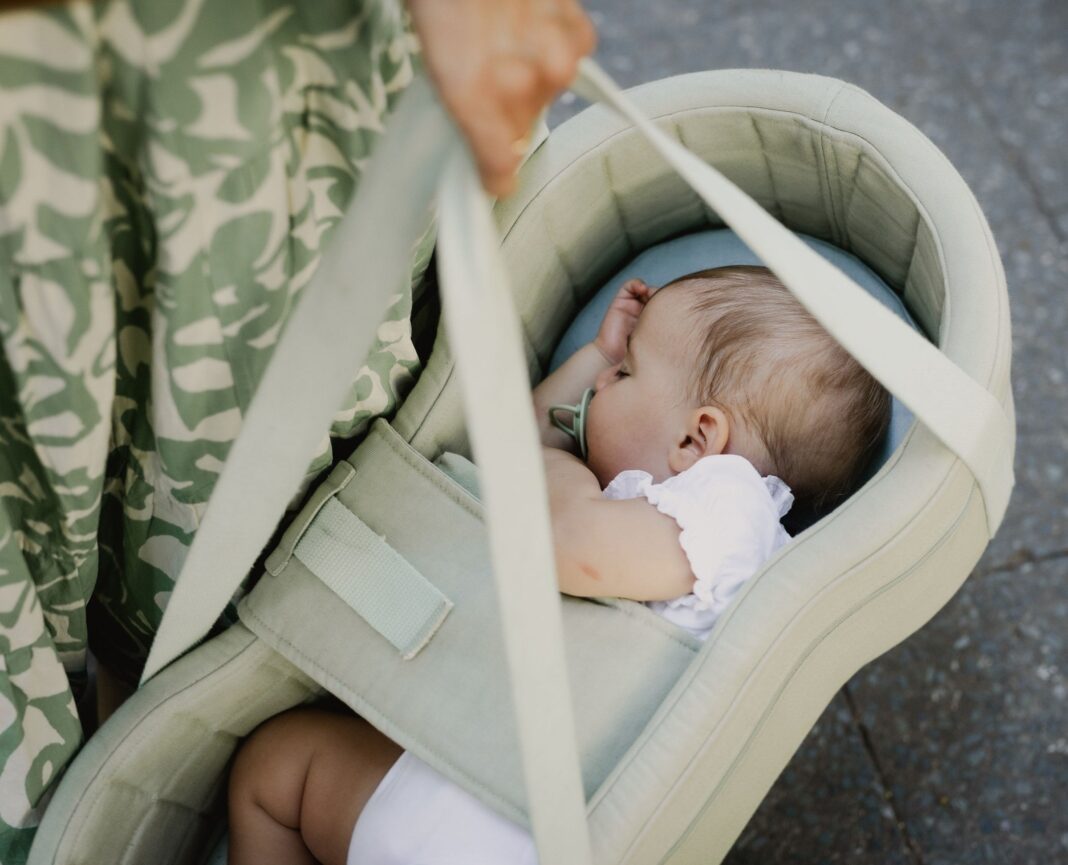 Ein schlafendes Baby liegt mit Schnuller seitlich in einer hellgruenen PEANUT-Tragetasche, die eine Person an zwei Griffen traegt