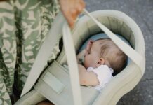 Ein schlafendes Baby liegt mit Schnuller seitlich in einer hellgruenen PEANUT-Tragetasche, die eine Person an zwei Griffen traegt