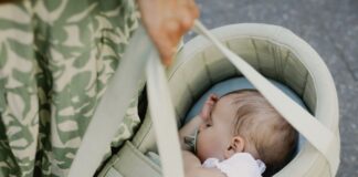 Ein schlafendes Baby liegt mit Schnuller seitlich in einer hellgruenen PEANUT-Tragetasche, die eine Person an zwei Griffen traegt