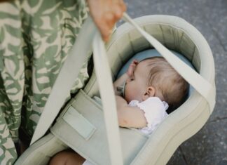 Ein schlafendes Baby liegt mit Schnuller seitlich in einer hellgruenen PEANUT-Tragetasche, die eine Person an zwei Griffen traegt