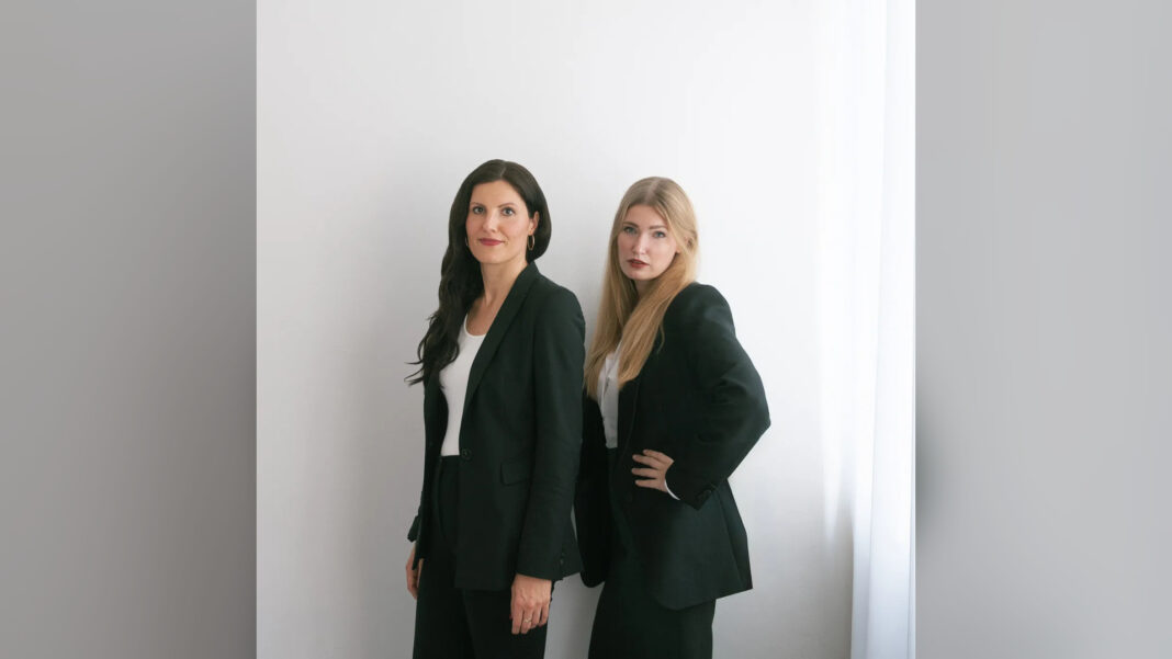 Die Gruenderinnen Carolin Krebber und Leonie Dowling stehen in dunklen Anzuegen nebeneinander vor einer hellen Wand