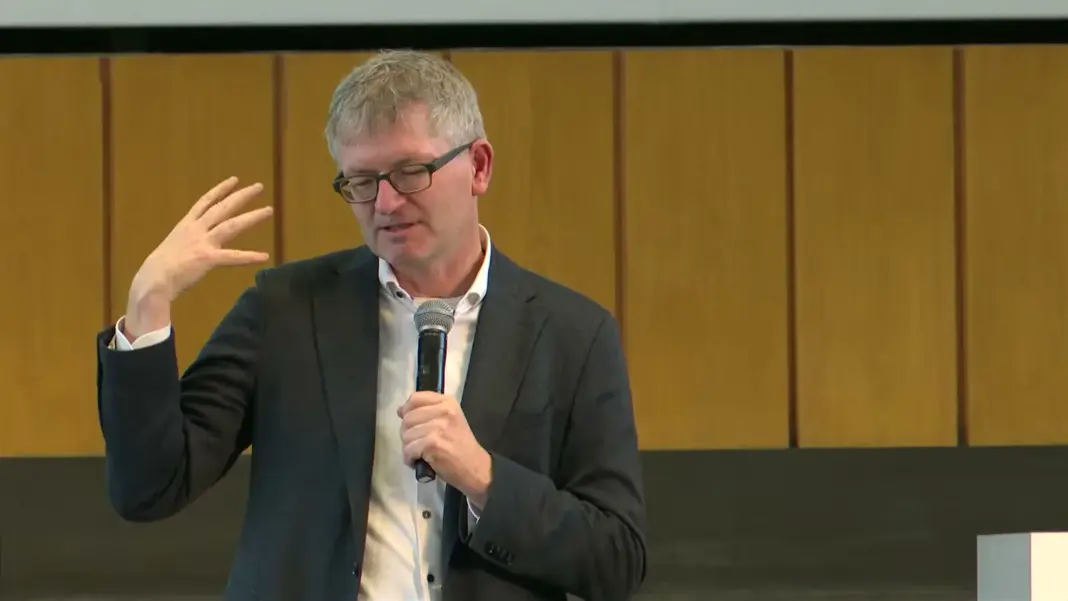Helmut Schönenberger von UnternehmerTUM spricht mit Mikrofon auf der Bühne im Audimax der FU Berlin vor der holzvertäfelten Wand