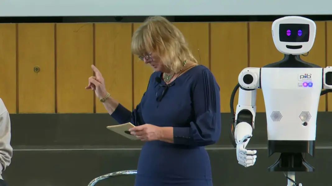 Moderatorin Tatjana Barzi-Vogt liest von einer Karte ab, neben ihr steht der weiße humanoide Roboter pib auf der Bühne