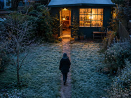 Ein blank getretener Pfad quert den bereiften Rasen vom Wohnhaus zur Tür eines kleinen Gartenateliers, aus dessen Sprossenfenster warmes Lampenlicht in die blaue Morgendämmerung fällt