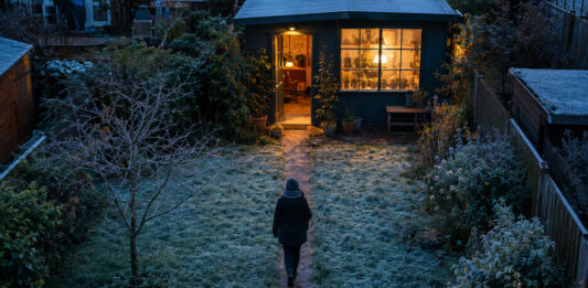 Ein blank getretener Pfad quert den bereiften Rasen vom Wohnhaus zur Tür eines kleinen Gartenateliers, aus dessen Sprossenfenster warmes Lampenlicht in die blaue Morgendämmerung fällt
