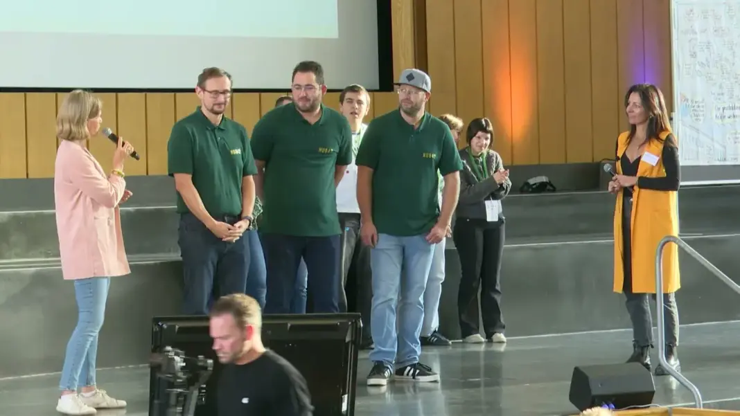 Drei Männer in grünen Poloshirts mit dem Aufdruck HUB42 stehen auf der Bühne des Audimax, der mittlere spricht ins Mikrofon