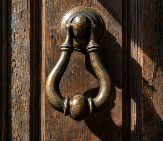 Ein schwerer Türklopfer aus Messing hängt an einer verwitterten Eichentür, das Metall dunkel patiniert, schräges Morgenlicht wirft einen harten Schatten auf das Holz.
