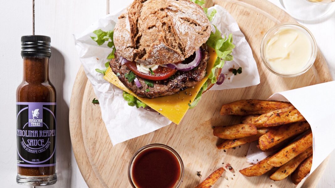 Burger mit Pommes auf einem Holzbrett, daneben eine Flasche Carolina Reaper Sauce von Mexican Tears und ein Schälchen Sauce
