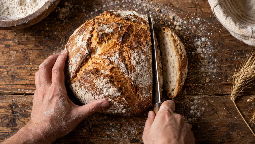 Ein breites Messer trennt ein schmales Stück von einem runden Brotlaib auf einem bemehlten Holztisch ab