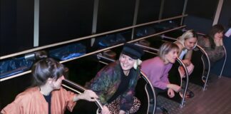 Fünf Frauen hocken nebeneinander an Missoir-Hockurinalen mit Haltegriffen in einer dunklen Toilettenanlage und lachen