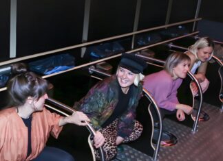 Fünf Frauen hocken nebeneinander an Missoir-Hockurinalen mit Haltegriffen in einer dunklen Toilettenanlage und lachen