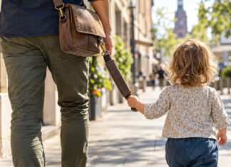 Braune Umhaengetasche mit gepolstertem Tragegriff, an dem sich ein kleines Kind auf dem Gehweg festhaelt