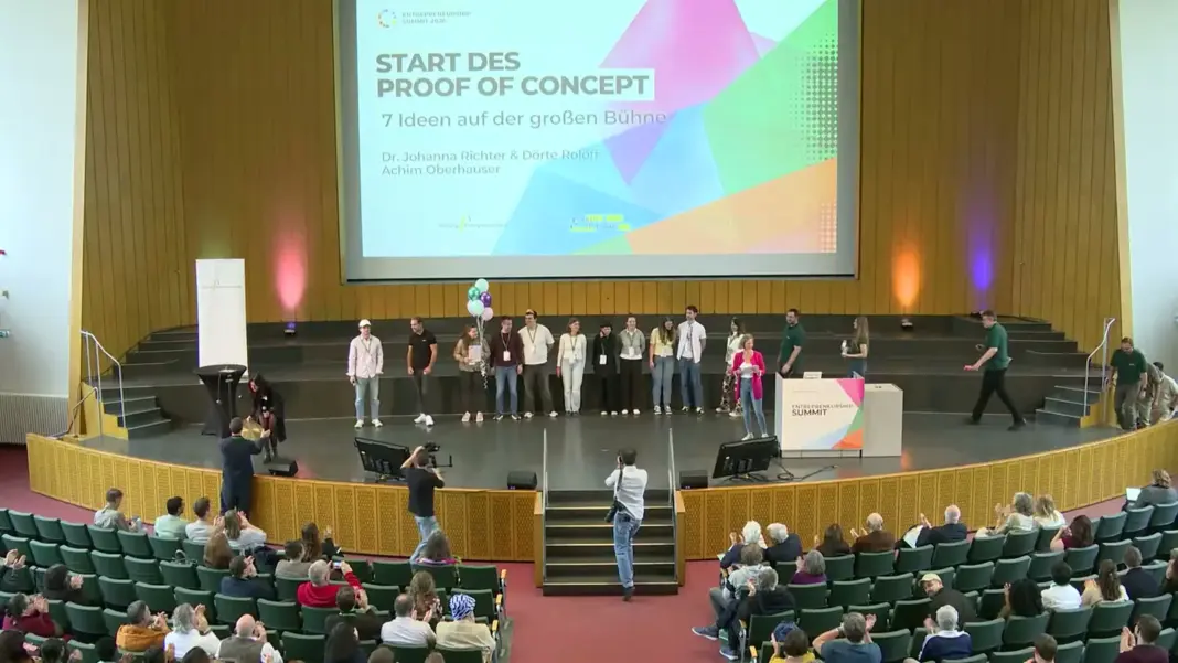 Blick in das Audimax beim Start des Proof of Concept: Die sieben Gründerteams stehen auf der Bühne, auf der Leinwand steht Start des Proof of Concept, 7 Ideen auf der großen Bühne