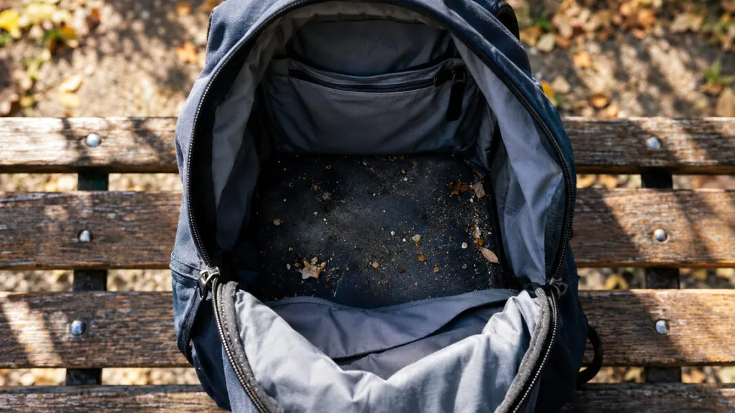 Offener Schulrucksack auf einer Parkbank mit Staub und Krümeln im Innenfach