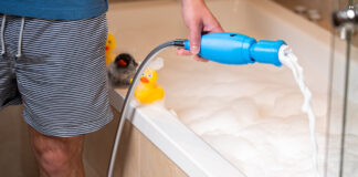 Ein Mann füllt mit dem blauen SchaumPo-Gerät am Duschschlauch Schaum in eine Badewanne, auf dem Wannenrand sitzen Quietscheenten