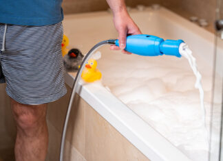 Ein Mann füllt mit dem blauen SchaumPo-Gerät am Duschschlauch Schaum in eine Badewanne, auf dem Wannenrand sitzen Quietscheenten