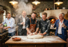 Fünf Männer stehen mehlbestäubt in einer rustikalen Küche um einen Pizzateig, im Hintergrund brennt ein Holzofen