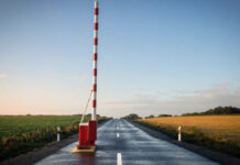 Ein rot-weiß gestreifter Schlagbaum steht senkrecht nach oben geöffnet über einer leeren Asphaltstraße, klares Morgenlicht auf dem frisch lackierten Holz und dem feuchten Asphalt