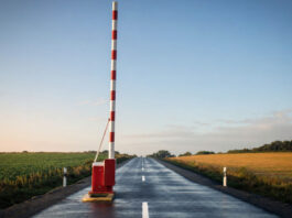 Ein rot-weiß gestreifter Schlagbaum steht senkrecht nach oben geöffnet über einer leeren Asphaltstraße, klares Morgenlicht auf dem frisch lackierten Holz und dem feuchten Asphalt