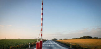 Ein rot-weiß gestreifter Schlagbaum steht senkrecht nach oben geöffnet über einer leeren Asphaltstraße, klares Morgenlicht auf dem frisch lackierten Holz und dem feuchten Asphalt