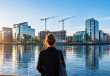 Eine Frau steht von hinten am Ufer der Liffey in Dublin, gegenüber Glasbürotürme und zwei Baukräne der Docklands im Morgenlicht (Symbolbild, mit KI erzeugt)