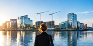 Eine Frau steht von hinten am Ufer der Liffey in Dublin, gegenüber Glasbürotürme und zwei Baukräne der Docklands im Morgenlicht (Symbolbild, mit KI erzeugt)