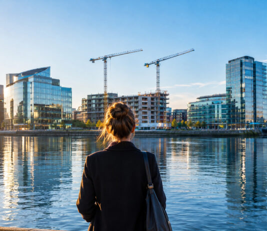 Eine Frau steht von hinten am Ufer der Liffey in Dublin, gegenüber Glasbürotürme und zwei Baukräne der Docklands im Morgenlicht (Symbolbild, mit KI erzeugt)