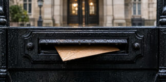 Ein schmaler brauner Umschlag steckt halb in einem gusseisernen Briefkastenschlitz, Regentropfen glänzen auf dem schwarz lackierten Metall