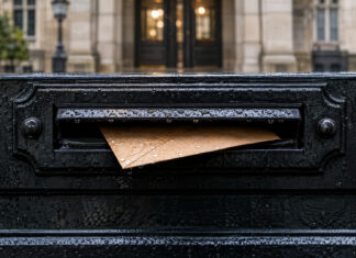Ein schmaler brauner Umschlag steckt halb in einem gusseisernen Briefkastenschlitz, Regentropfen glänzen auf dem schwarz lackierten Metall