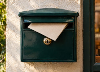 Ein cremefarbener Leinenumschlag steckt zur Hälfte in der Klappe eines emaillierten Wandbriefkastens, das Messingschloss blank gegriffen, weiches Morgenlicht von der Seite